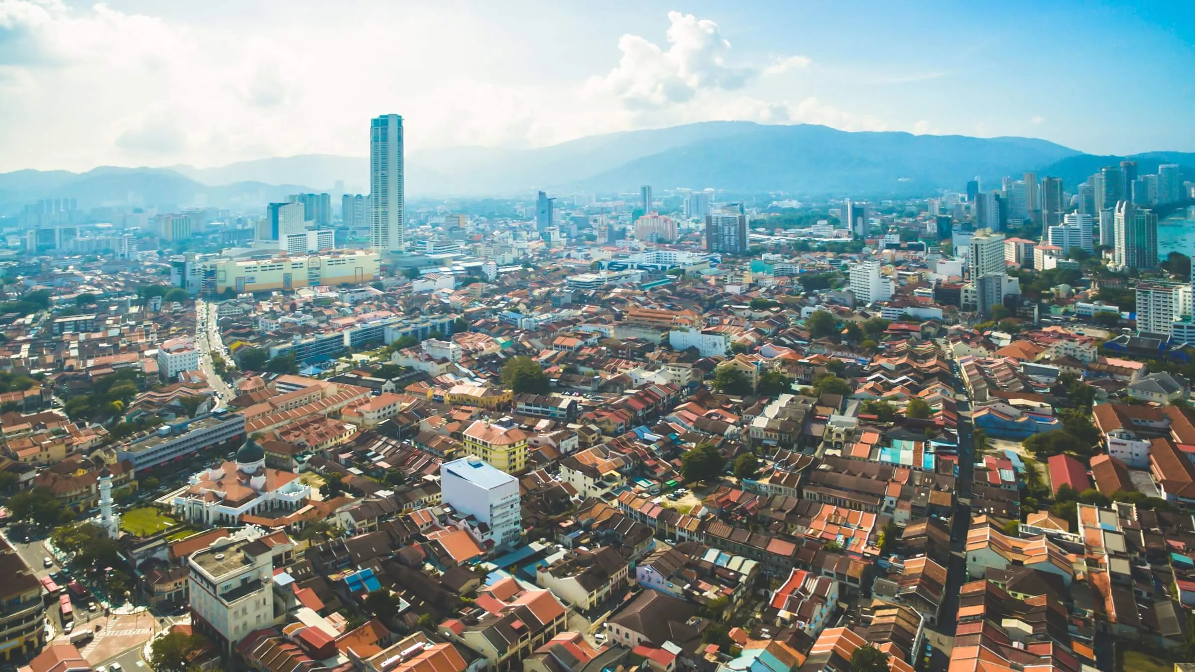 Aerial view over the red-tiled shophouse roofs of George Town's old town, with the KOMTAR tower rising above the city and the hills of Penang behind